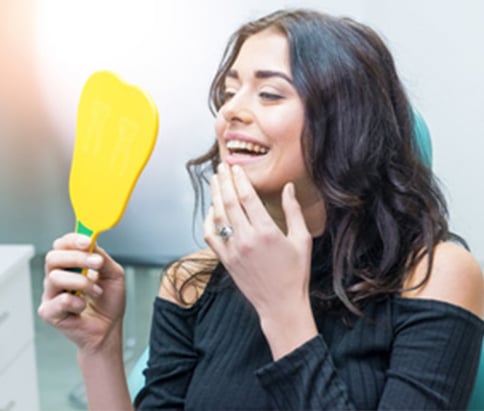 Beautiful young woman checking new dental implants in yellow hand mirror