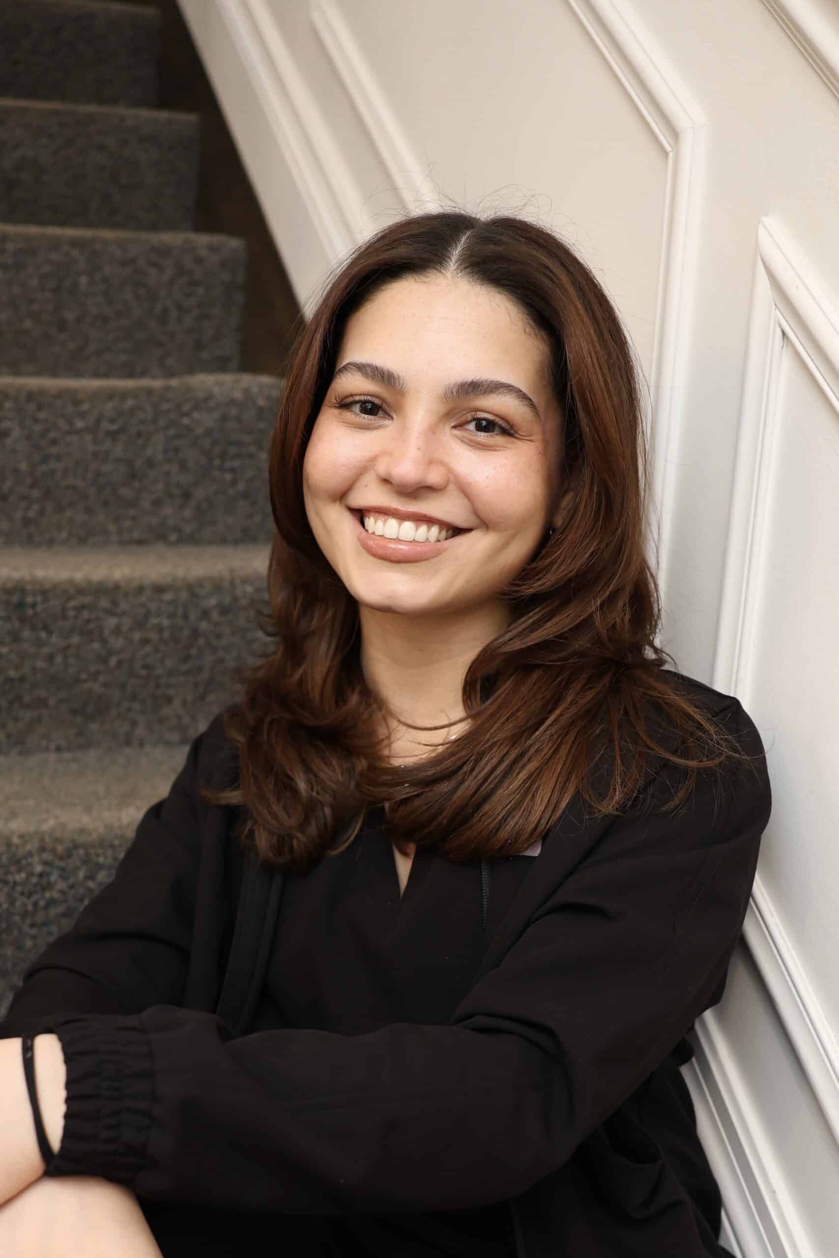 A woman with brown hair and a black top sits on carpeted stairs, smiling at the camera with a white wall in the background.