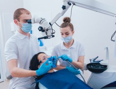 Two dental professionals operate on a patient seated in a dental chair, performing a root canal procedure with various dental instruments and wearing protective masks and gloves.